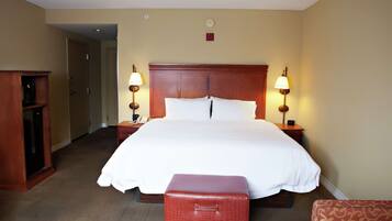 A hotel room with a large bed, two bedside tables with lamps, a wooden headboard, a small red suitcase, and a carpeted floor.