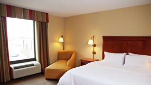 Habitación de hotel con cama, una silla, una mesita de noche, lámpara, equipo de aire acondicionado y ventana con cortinas.