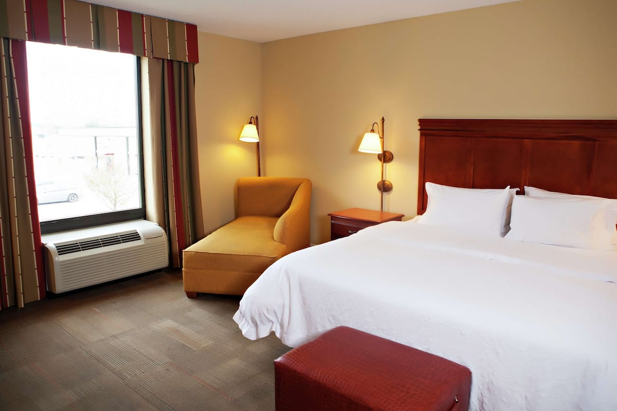 A hotel room with a bed, a chair, a nightstand, a lamp, an air conditioning unit, and a window with curtains.