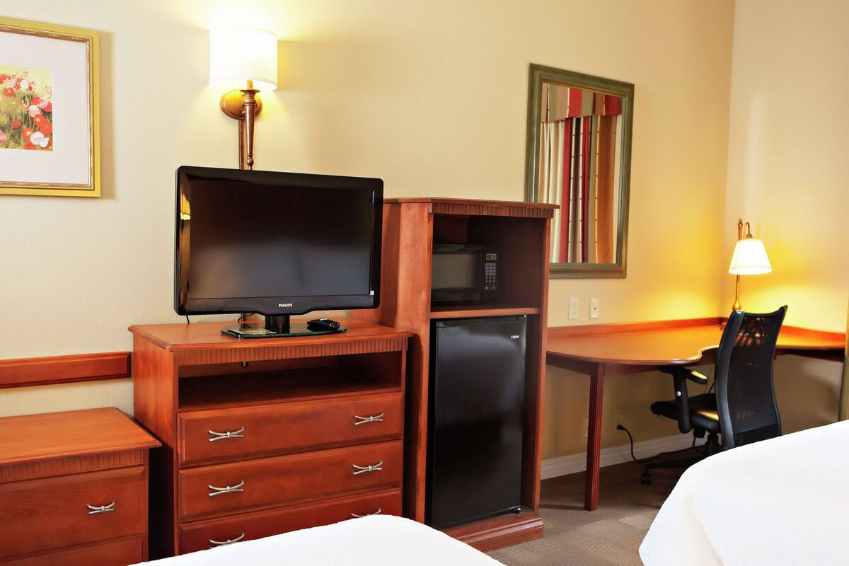 A hotel room with a bed, a desk with a computer, a refrigerator, a chair, and a lamp.