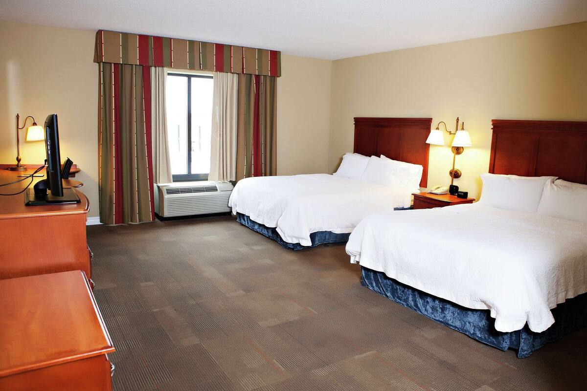 A hotel room with two beds, a desk, a chair, an air conditioning unit, and a window with curtains.