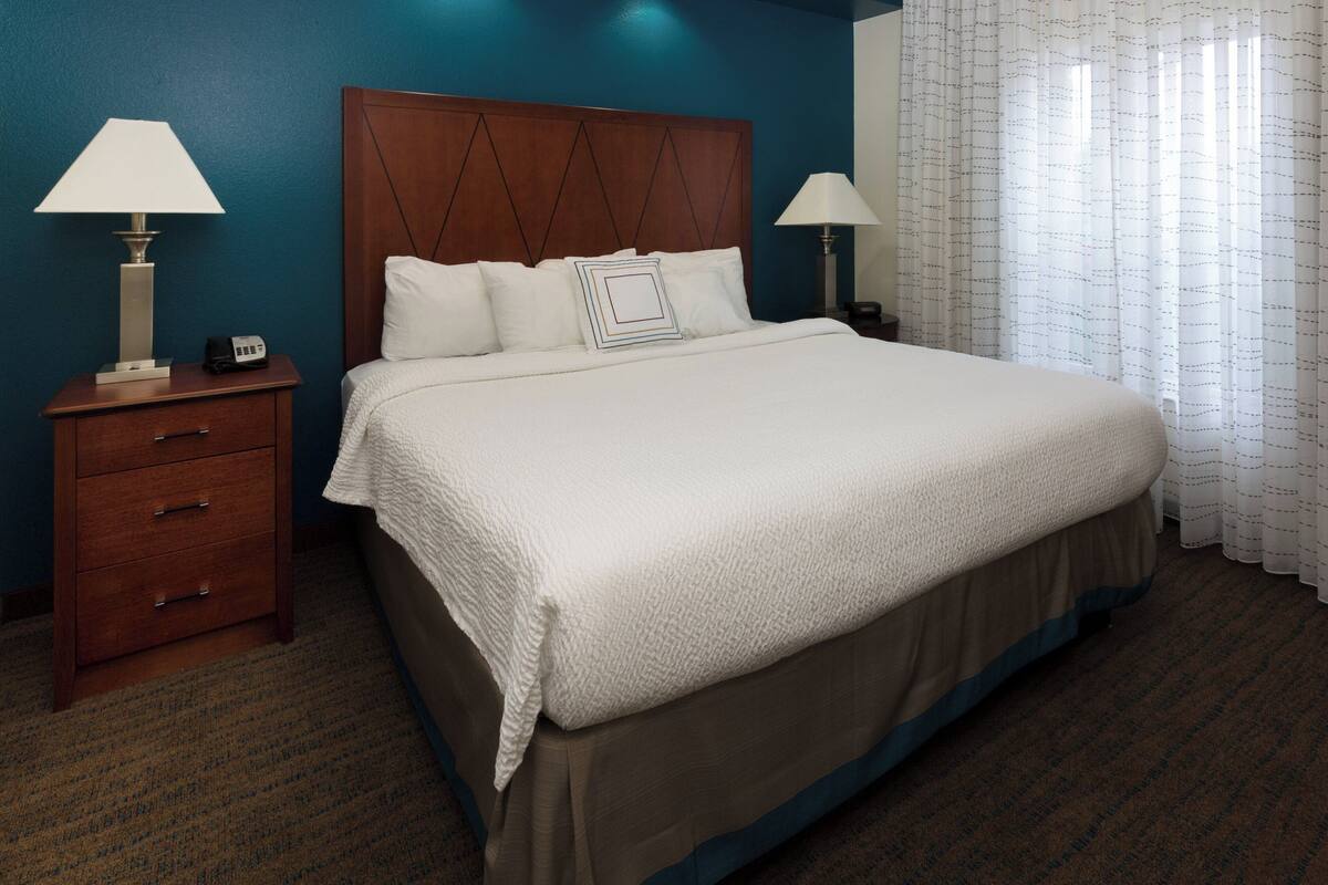A neatly made bed with a white comforter and a wooden headboard, flanked by two bedside tables with lamps, one of which has a phone on it.