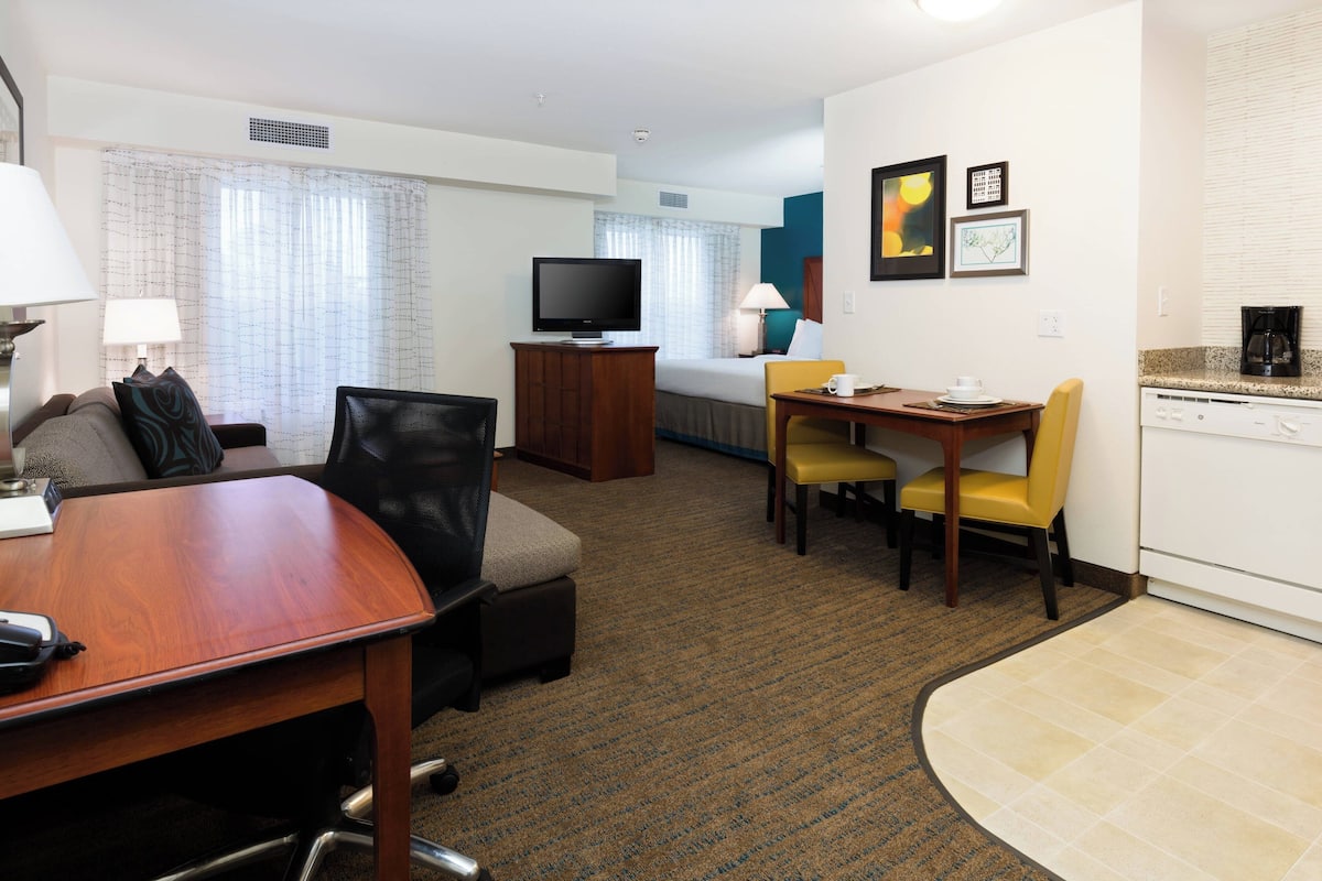 A hotel room with a wooden desk, a television, a bed, a dining table with chairs, and a kitchen area.