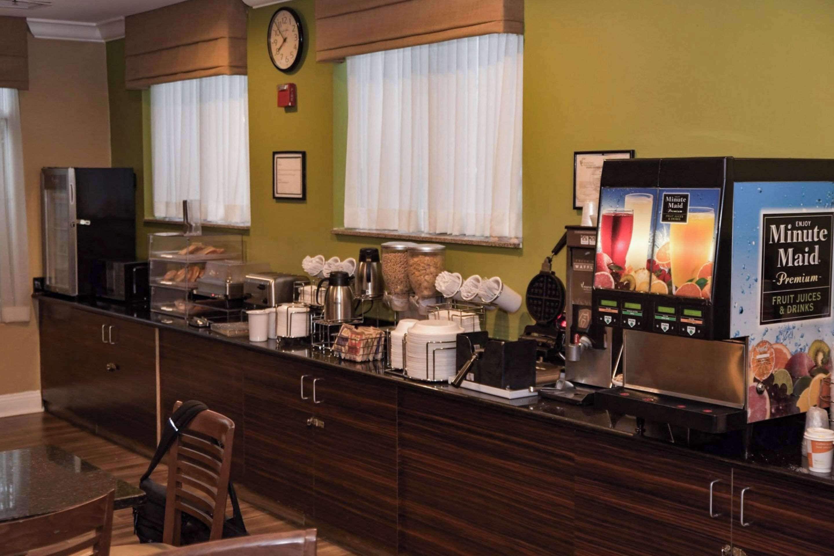 A breakfast bar with a coffee machine, toaster, and various food items. There is a clock on the wall and a window with blinds.