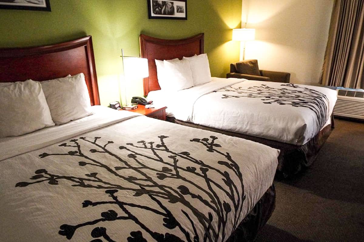 A hotel room with two beds, each with a floral-patterned bedspread, a wooden headboard, a bedside table with a lamp, and a chair.