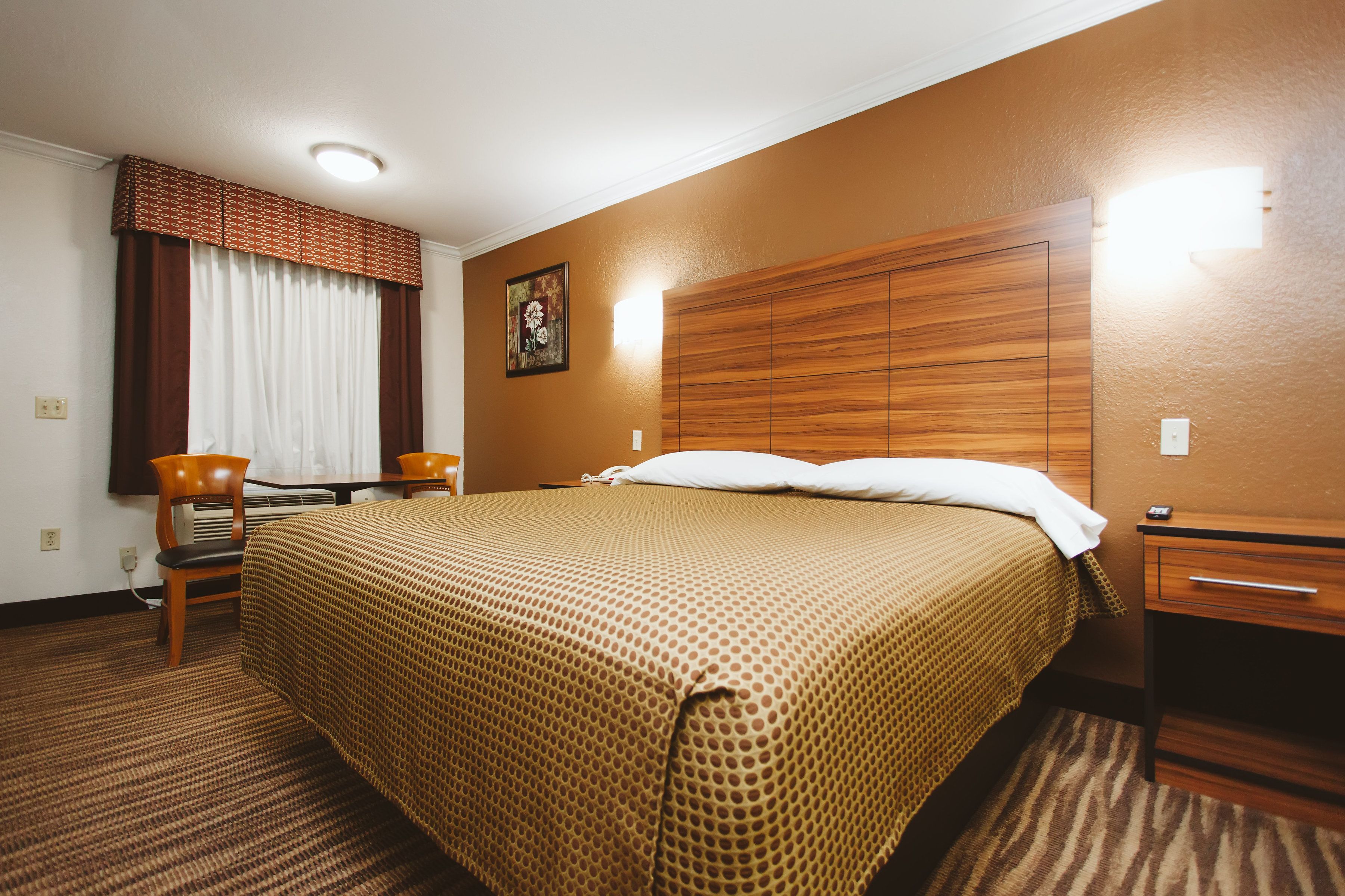 Habitación de hotel con una cama grande, cabecera de madera, escritorio con silla, mesita de noche y ventana con cortinas.