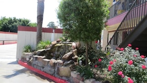 A landscaped area with a stone retaining wall, a palm tree, and flowering plants. - Palace Inn Wayside (Houston)