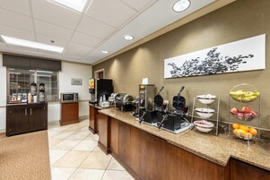 Zona de desayuno de hotel con mostrador, máquinas de café, microondas y una exposición de frutas.