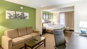Habitación de hotel con pared verde, sofá color beige, una mesita pequeña, un escritorio con silla, cama con cabecera, dos mesitas de noche con lámparas y una ventana con cortinas.