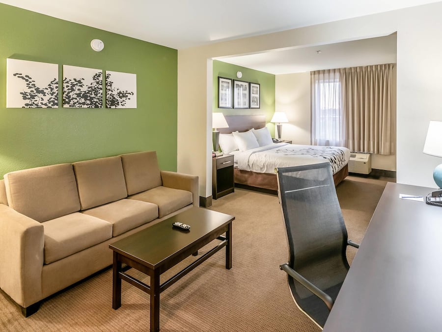 A hotel room with a green wall, a beige sofa, a small table, a desk with a chair, a bed with a headboard, two bedside tables with lamps, and a window with curtains.