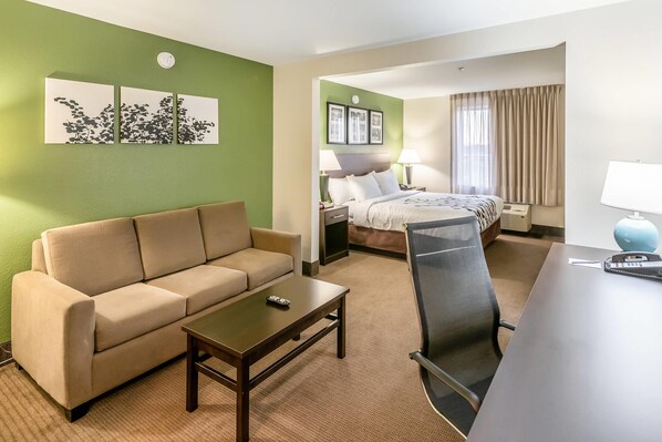 A hotel room with a green wall, a beige sofa, a small table, a desk with a chair, a bed with a headboard, two bedside tables with lamps, and a window with curtains. - Sleep Inn & Suites Ruston Near University (Ruston)