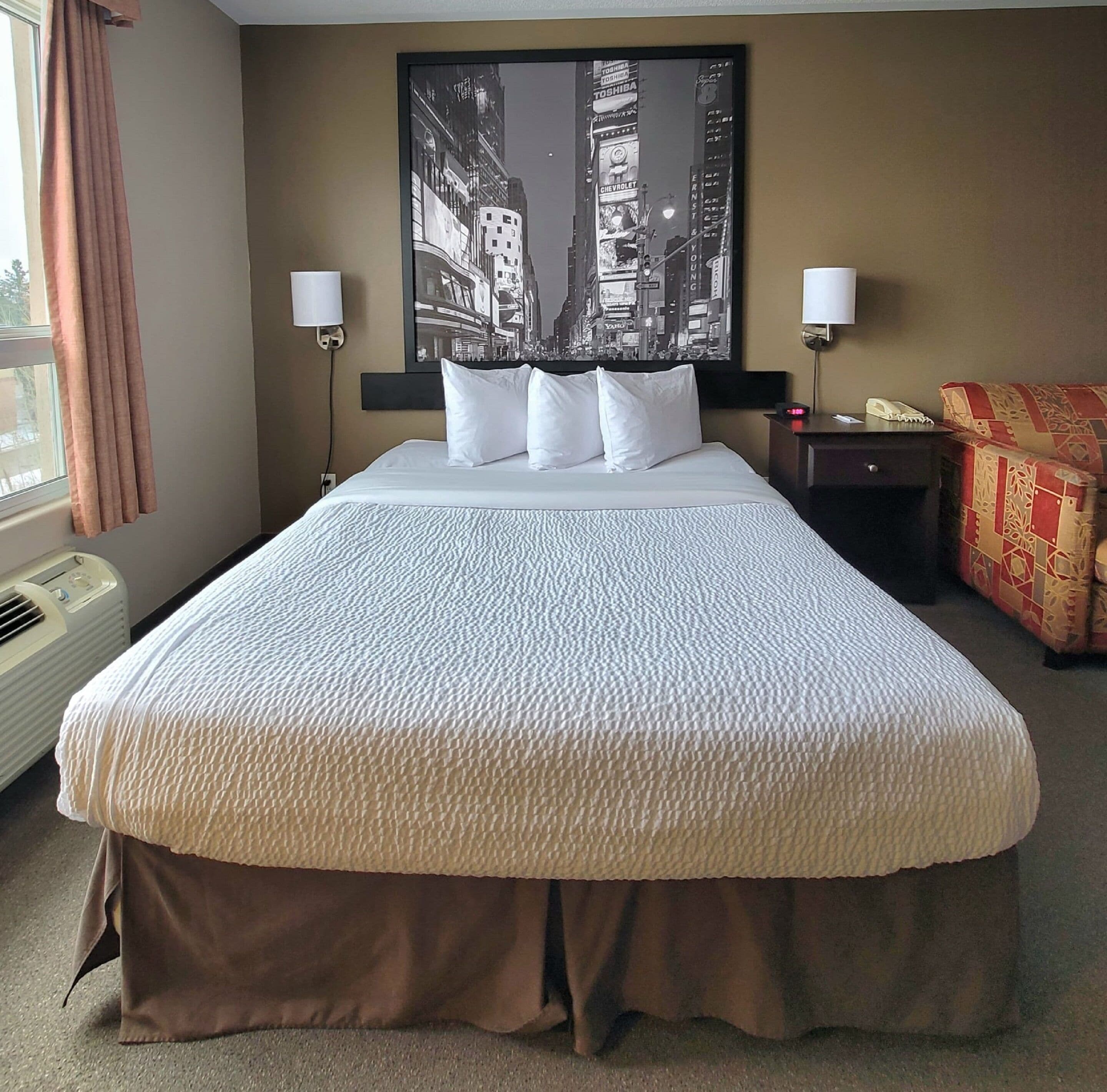 A hotel room with a large bed, two bedside lamps, a nightstand, and a cityscape painting on the wall.