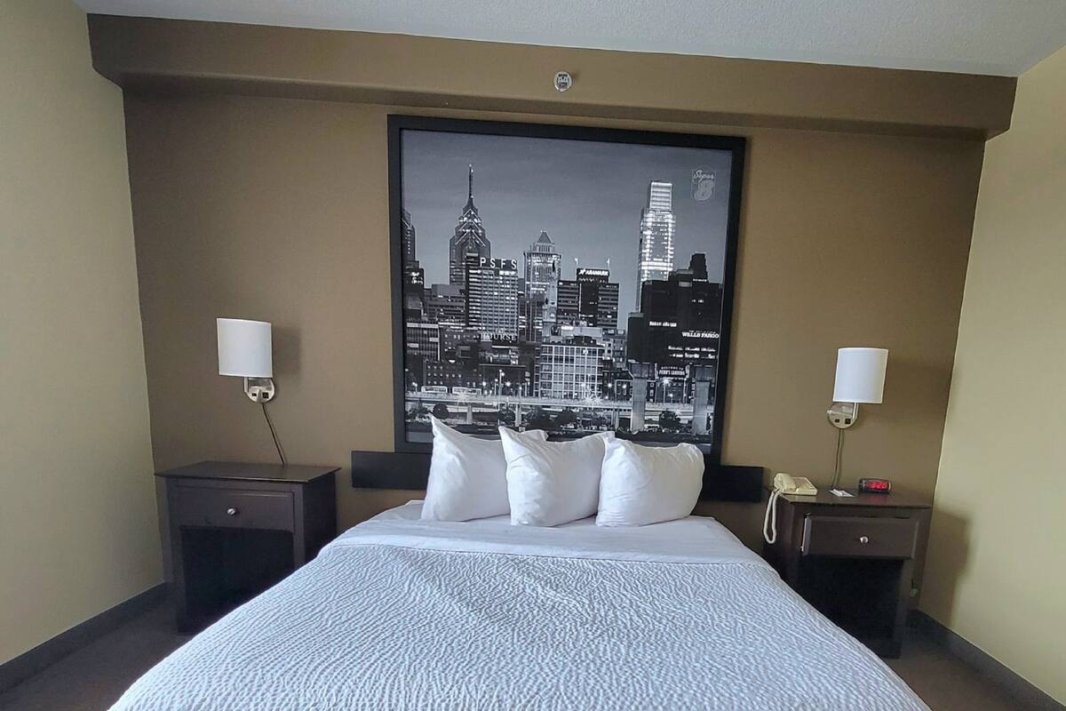 Une chambre d’hôtel avec un grand lit, deux tables de chevet, une petite table d’appoint, une affiche représentant un paysage urbain accrochée au mur et deux lampes.