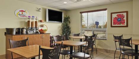 Cafetería para desayunos con mesas de madera y sillas negras, una máquina de café y un televisor montado en la pared.