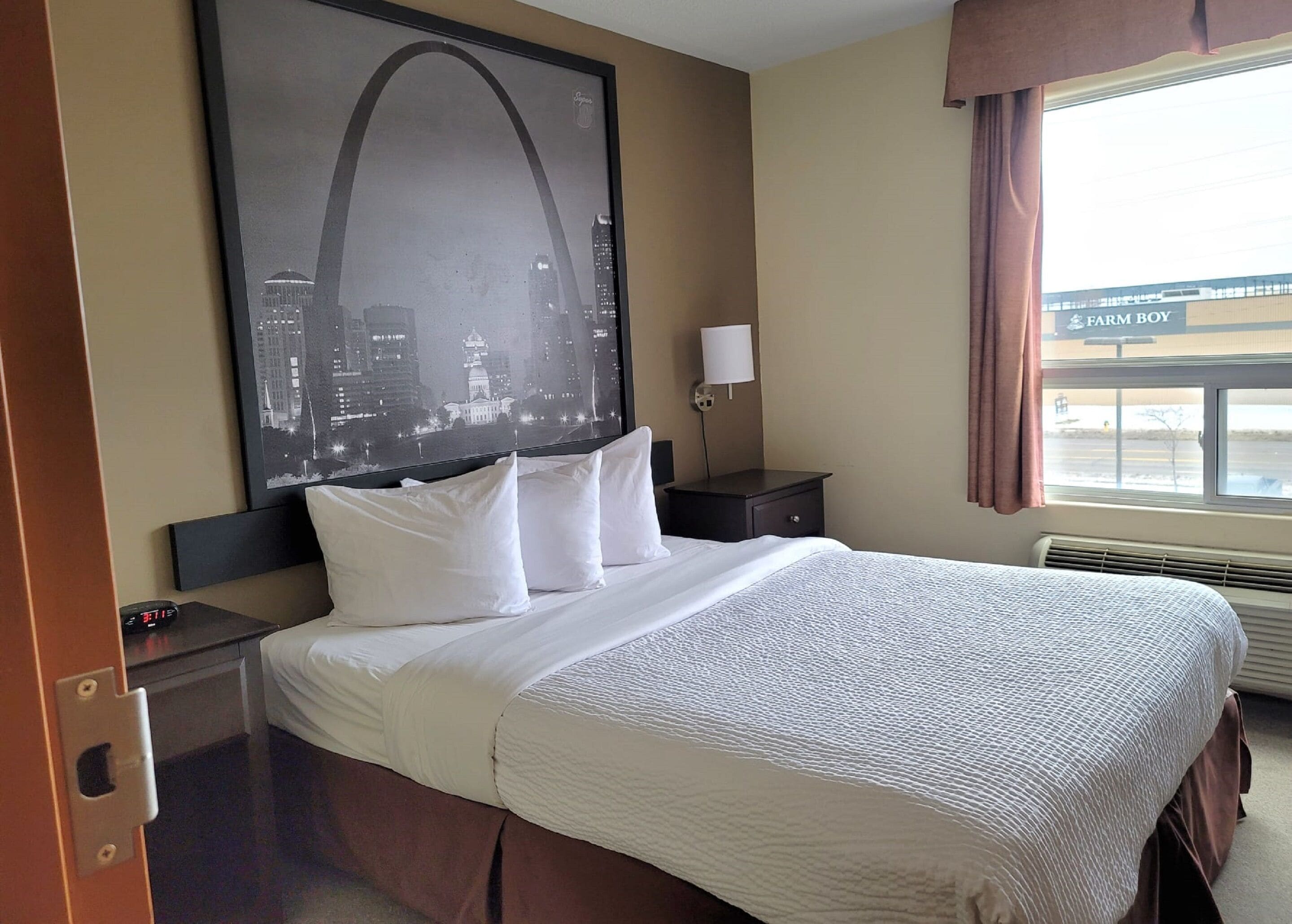 A hotel room with a large bed, a nightstand, a lamp, a window with a view of a building, and a framed picture on the wall.