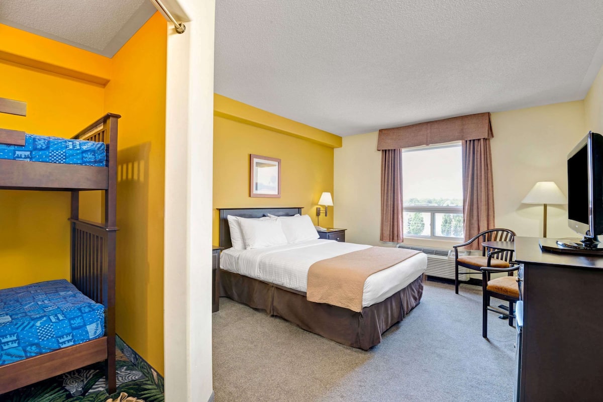 Une chambre d’hôtel avec un lit superposé, un lit simple, un bureau avec une chaise, une télévision, une fenêtre avec des rideaux et un tableau encadré au mur.