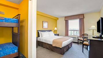 A hotel room with a bunk bed, a single bed, a desk with a chair, a TV, a window with curtains, and a framed picture on the wall.