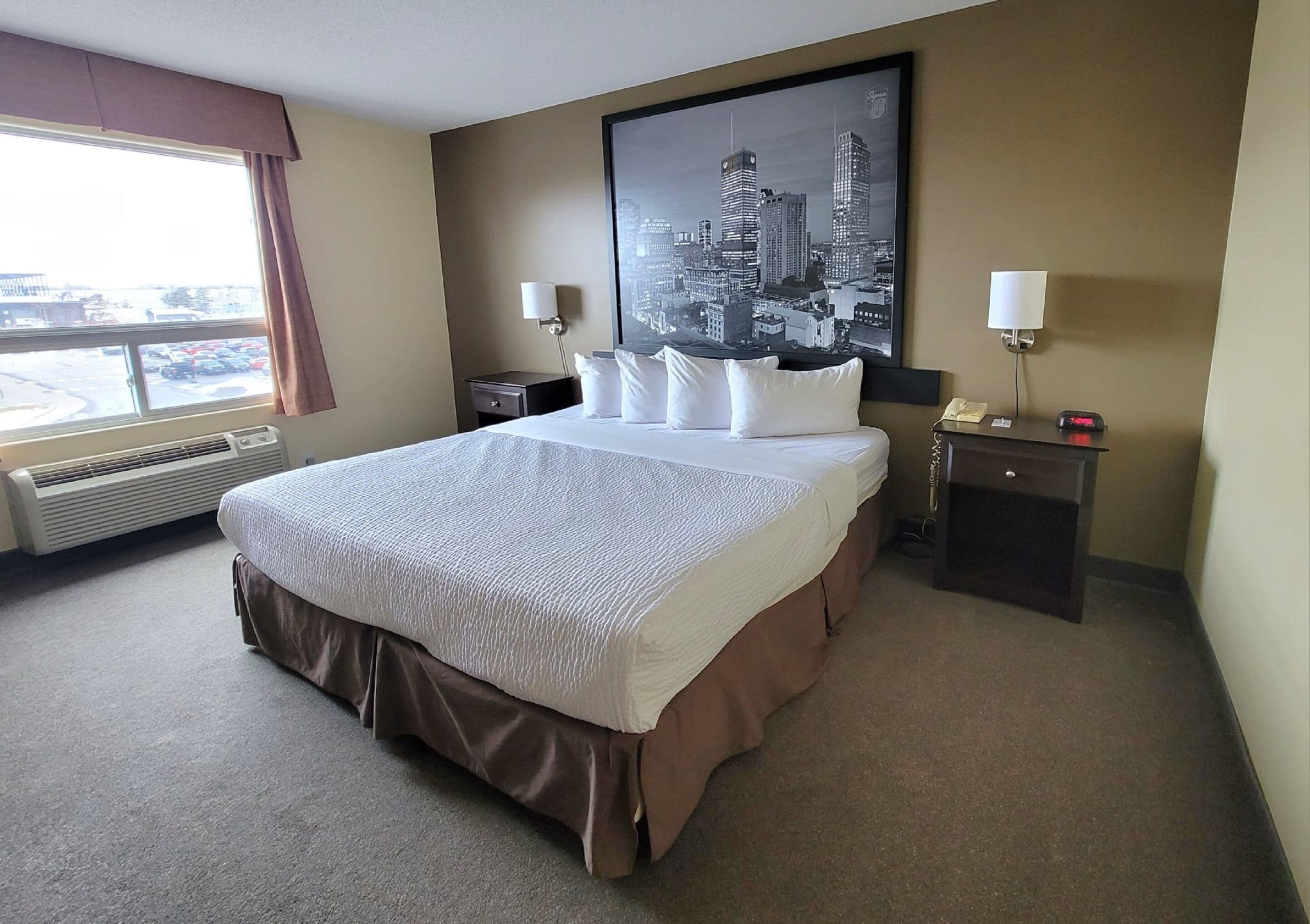 A hotel room with a large bed, a nightstand, a desk, a window with a view of the city, and a cityscape painting on the wall.
