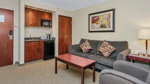 A compact living space with a kitchenette, a sofa with patterned cushions, a small table, and a lamp. - Holiday Inn Hotel & Suites Lloydminster by IHG (Lloydminster)