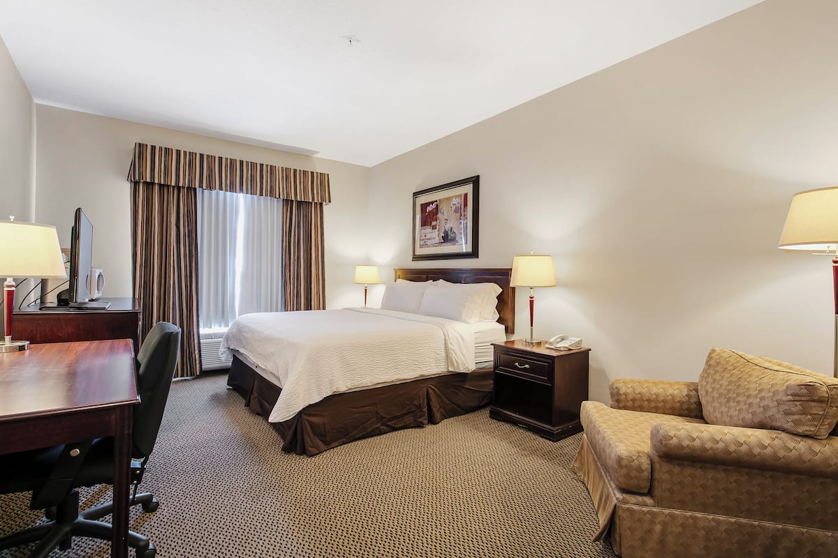 Une chambre d’hôtel comprenant un lit, deux tables de chevet avec des lampes, un bureau avec une chaise, un canapé et une grande fenêtre avec des rideaux.