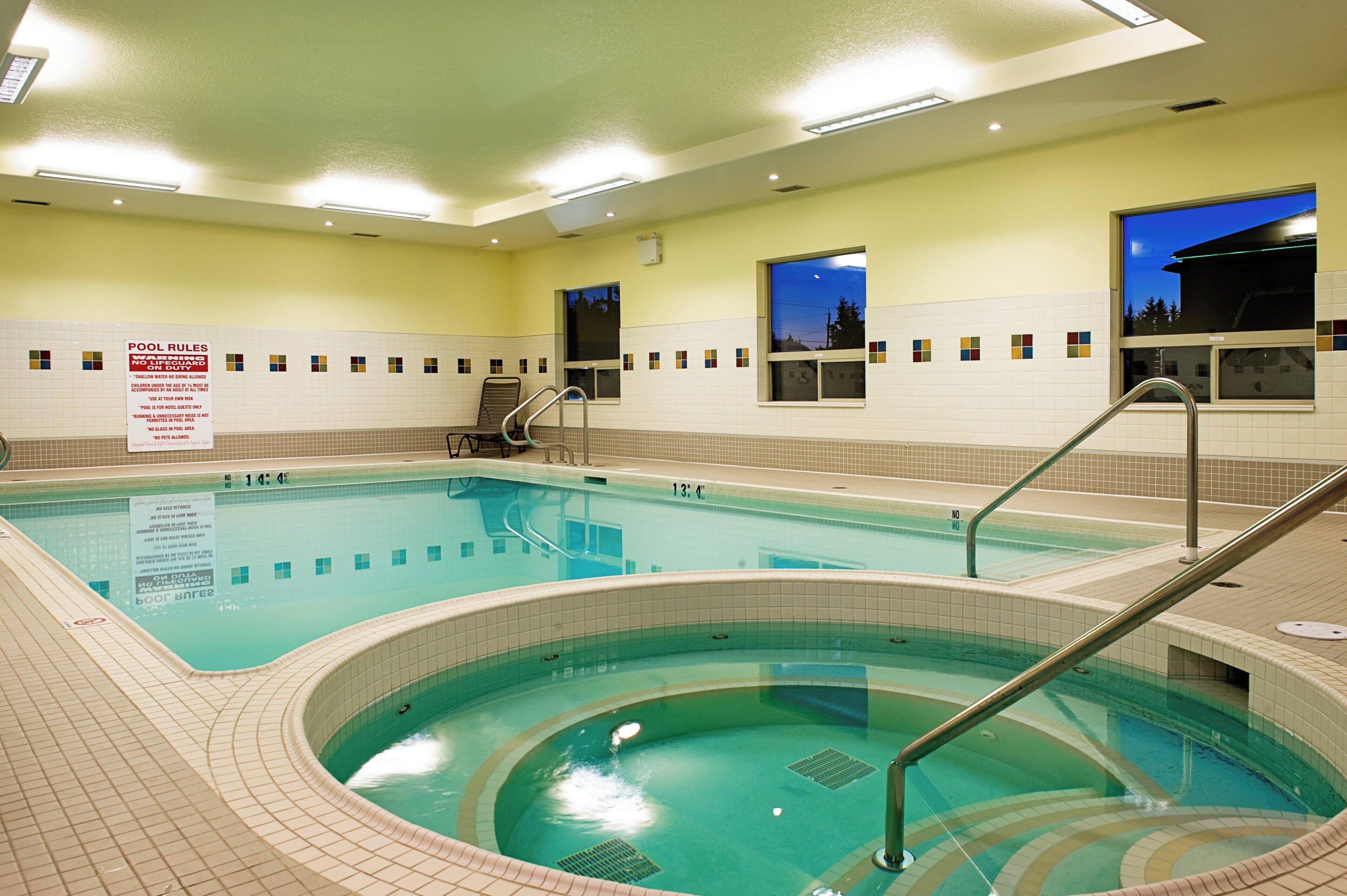An indoor swimming pool with a hot tub, tiled walls, and a pool rule sign.