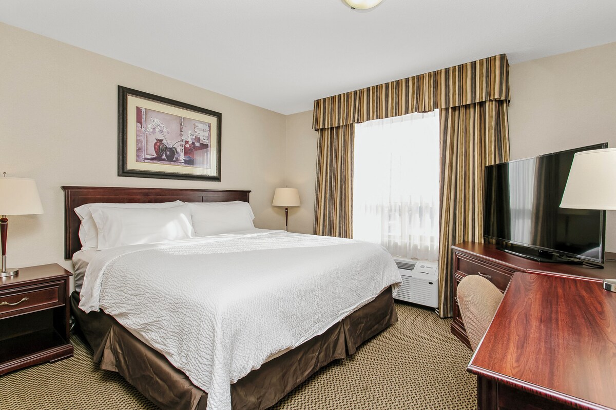 A hotel room with a large bed, a desk with a television, a framed picture on the wall, and a window with curtains.