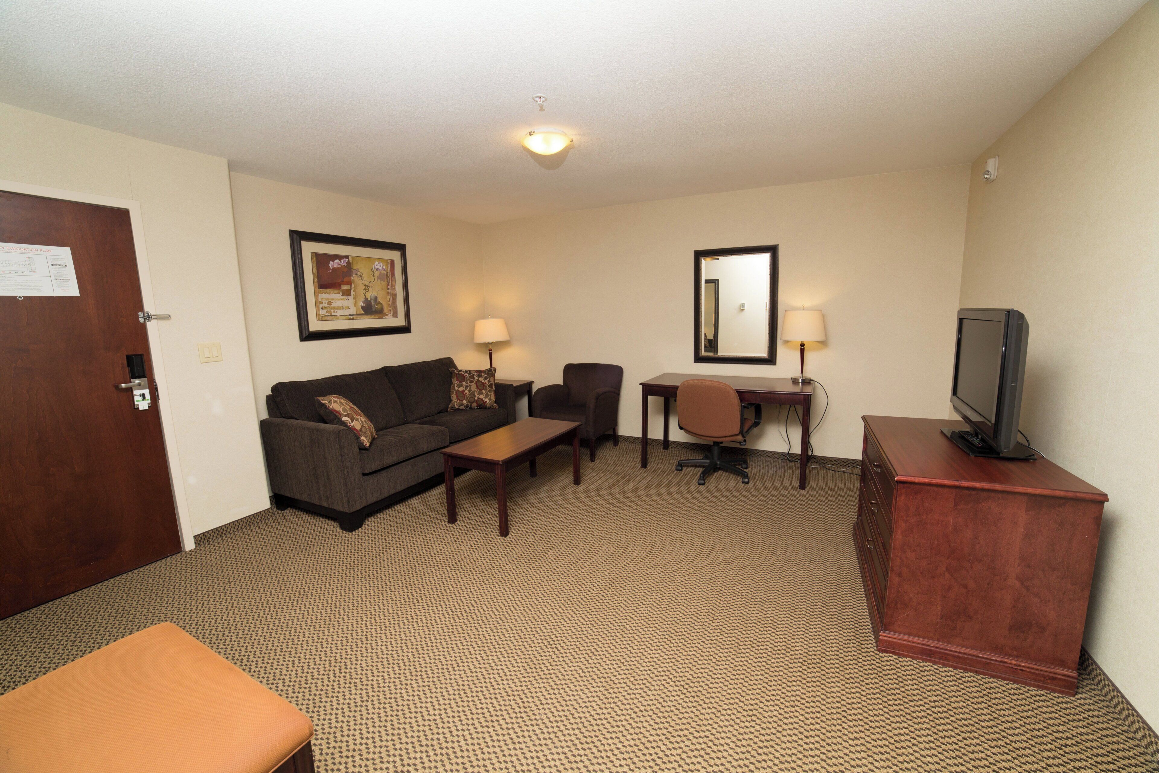 A hotel room with a sofa, a small table, a desk with a chair, a television on a cabinet, and a mirror on the wall.