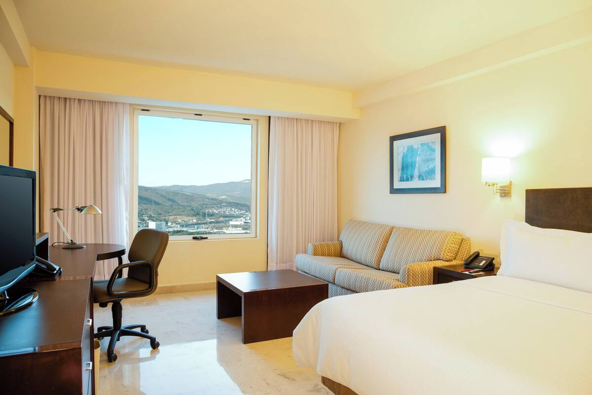 A hotel room with a bed, a desk with a computer, a chair, a sofa, and a view of the city.