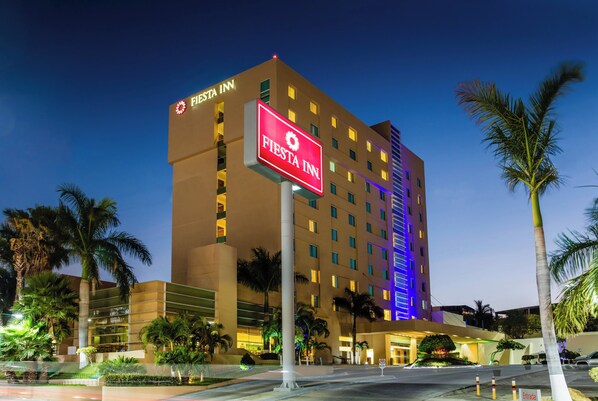 A multi-story hotel with a prominent 'Fiesta Inn' sign, palm trees, and a well-lit entrance area. - Fiesta Inn Tuxtla (Tuxtla Gutierrez)