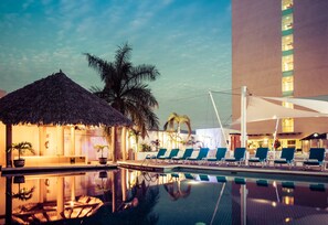 A pool area with lounge chairs and a thatched structure, surrounded by palm trees and a modern building.