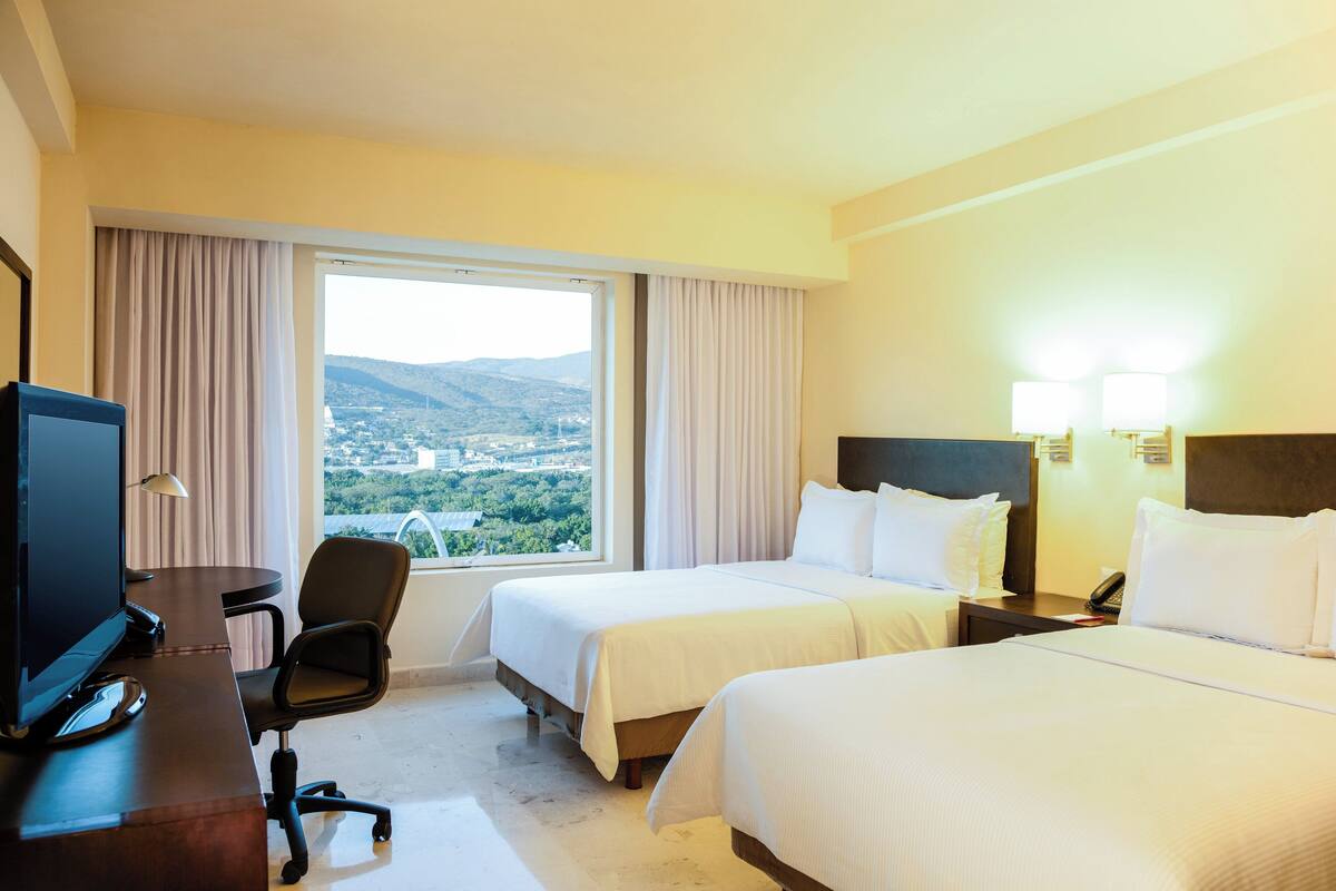 A hotel room with two beds, a desk with a computer, a chair, and a view of a city and mountains.
