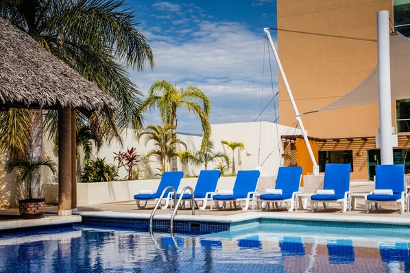 Piscina com espreguiçadeiras azuis, palmeiras e uma estrutura de telhado de palha.