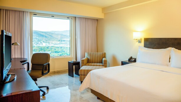 A hotel room with a bed, a desk with a chair, a TV, a lamp, a nightstand with a phone, and a view of the outdoors.