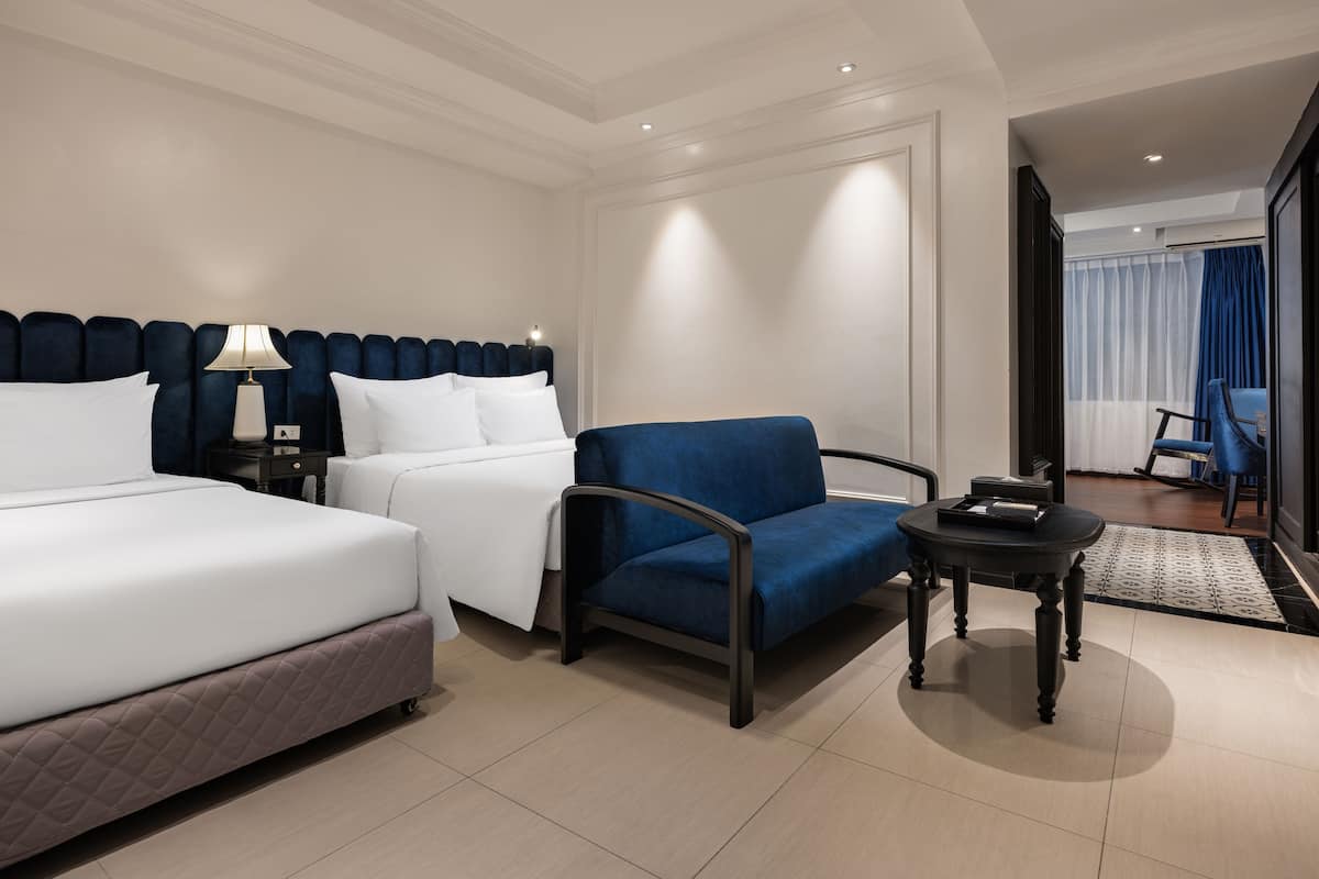 Habitación de hotel con dos camas, un sillón azul, una mesita y vista a la habitación contigua.