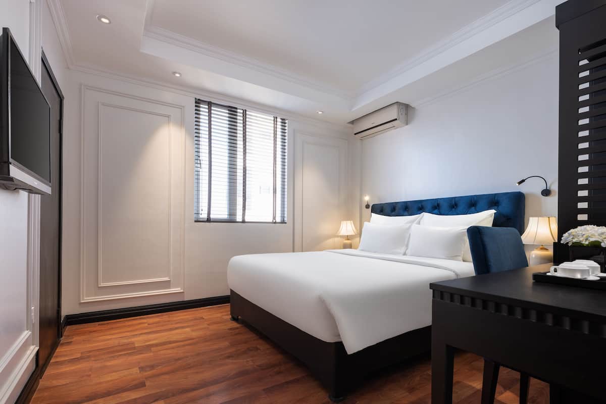 Habitación de hotel con una cama grande, una mesita de noche, un televisor montado en la pared y una ventana con persianas.
