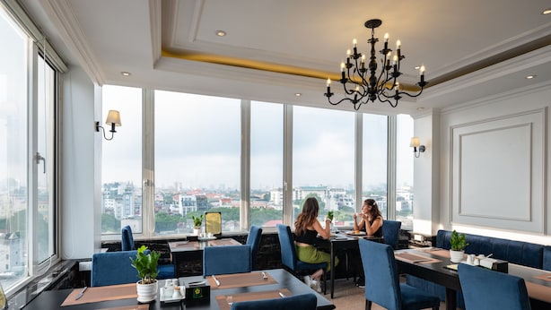 A restaurant with high ceilings, large windows, and a city view. Tables are set with blue chairs, and a chandelier hangs from the ceiling.
