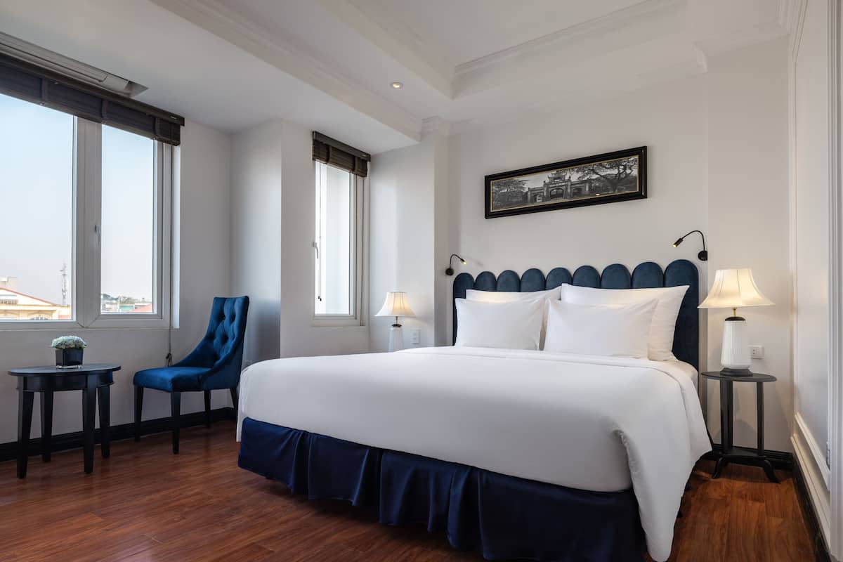 Habitación de hotel con una cama grande, dos mesitas de noche, una silla y una ventana con vistas.