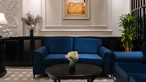 A modern lobby with a blue sofa set, a round coffee table with a vase of flowers, a framed artwork on the wall, and a potted plant.