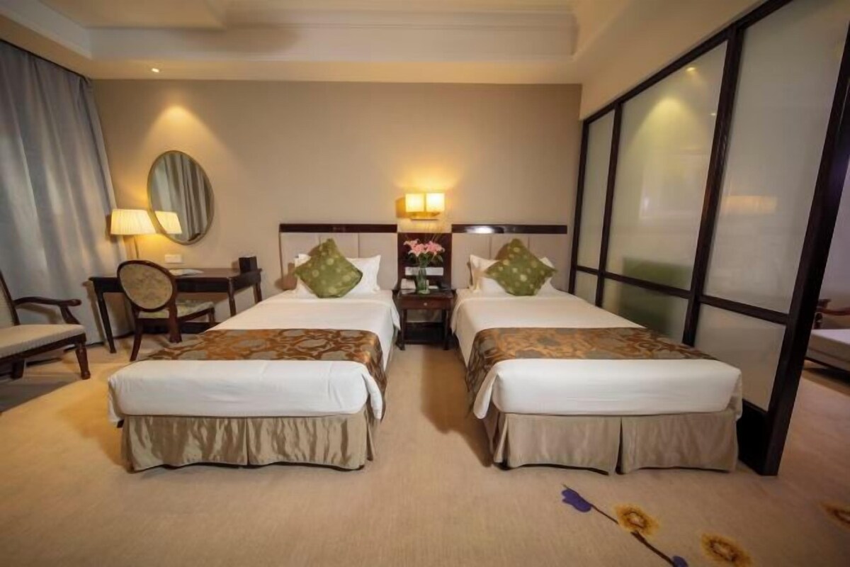 Quarto de hotel com duas camas, uma escrivaninha, uma cadeira, um abajur e uma janela grande.