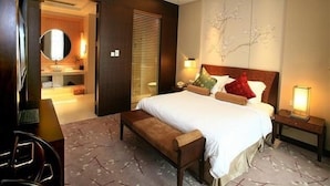 Une chambre d’hôtel avec un grand lit, une banquette, une table de chevet avec une lampe, un miroir fixé au mur et une vue sur la salle de bain.