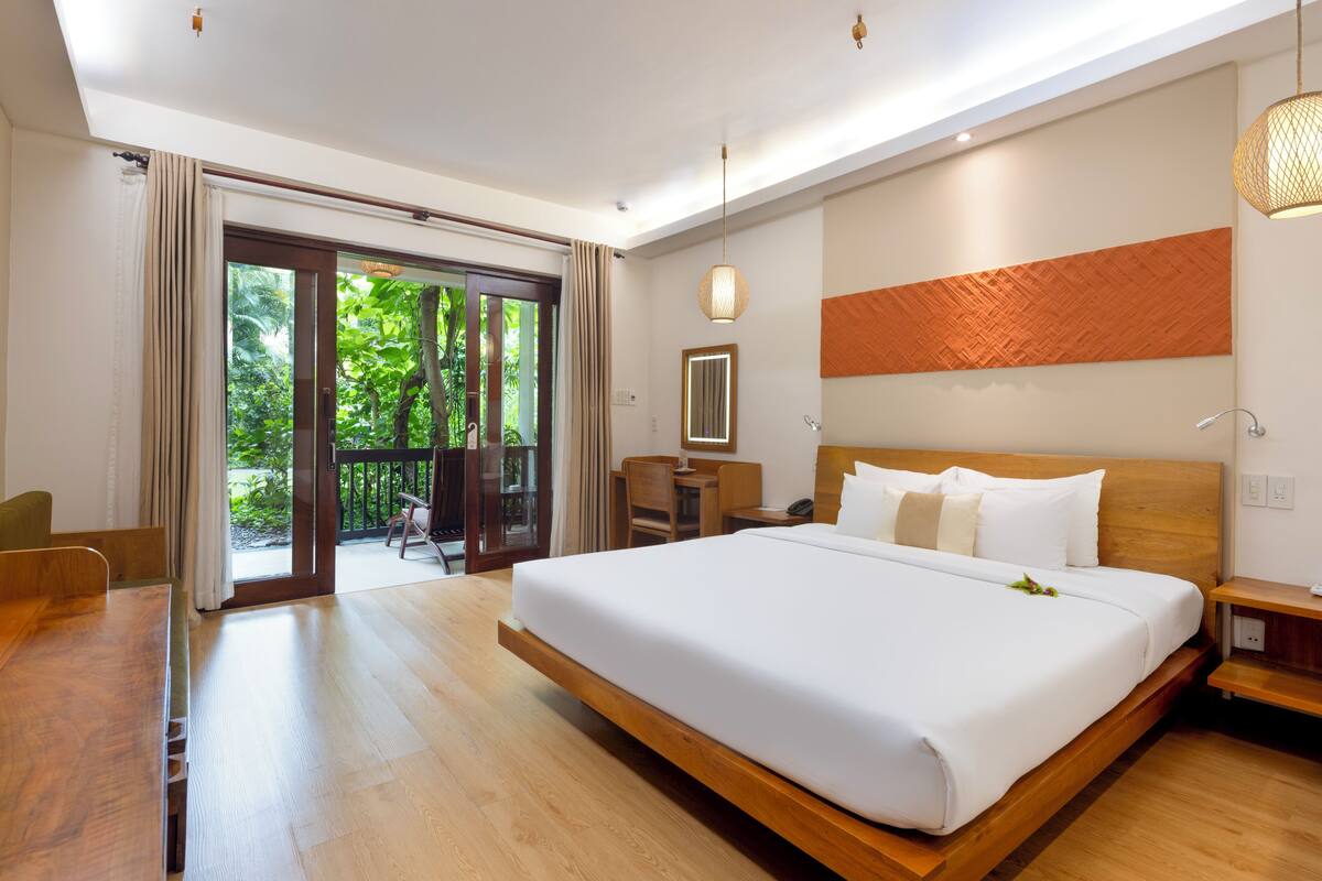 A modern hotel room with a large bed, wooden flooring, a desk, and a view of lush greenery through the sliding glass door.
