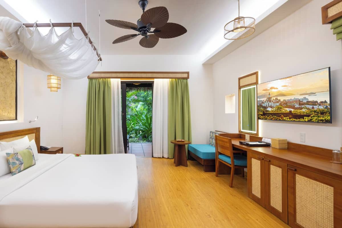 A hotel room with a large bed, a desk, a chair, a television, and a view of greenery outside.