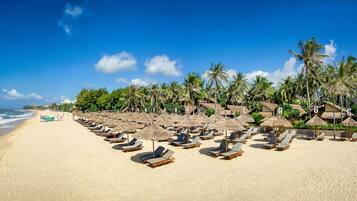 A beach with rows of sun loungers and thatched umbrellas, palm trees, and beach huts.