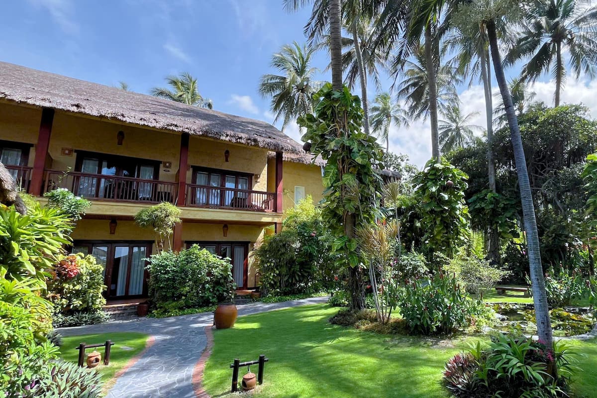 A two-story building with a thatched roof, surrounded by lush greenery and palm trees.