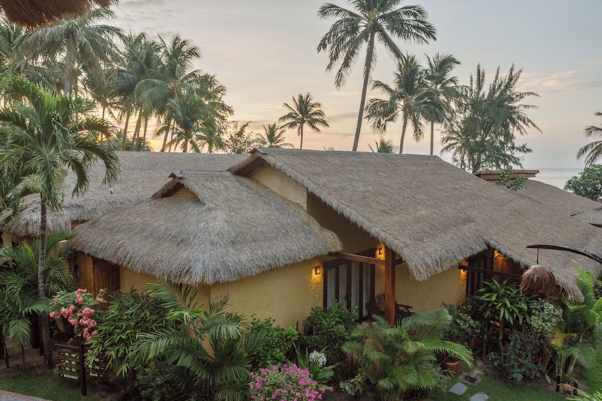 A thatched-roof bungalow with a garden, surrounded by palm trees.
