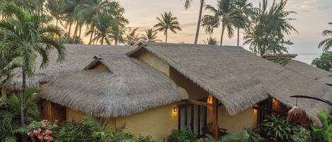 A thatched-roof bungalow with a garden, surrounded by palm trees.