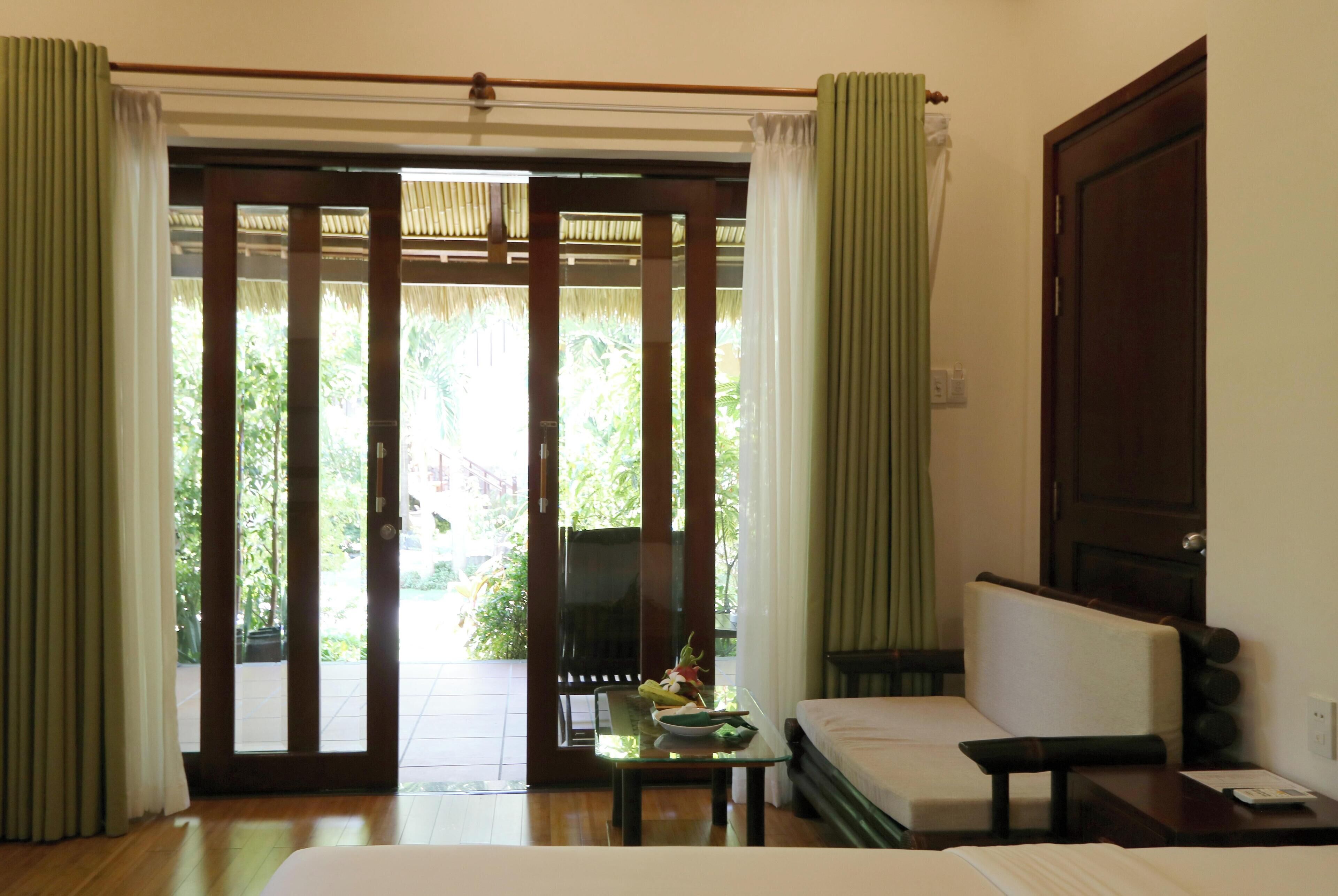 Habitación de hotel con cama, sofá, mesa con frutero y vistas a zonas verdes a través de una puerta corredera.