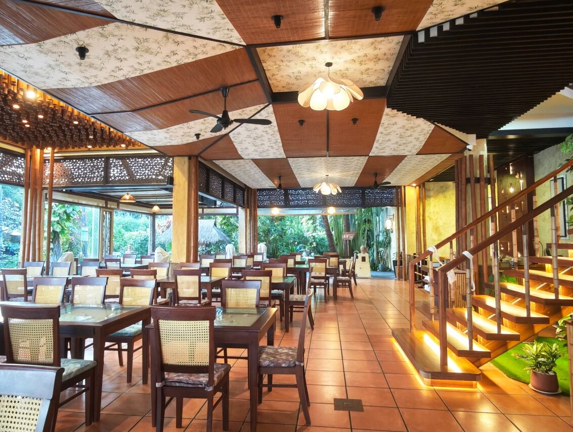 A restaurant with wooden tables and chairs, a staircase, and a ceiling with a geometric pattern.