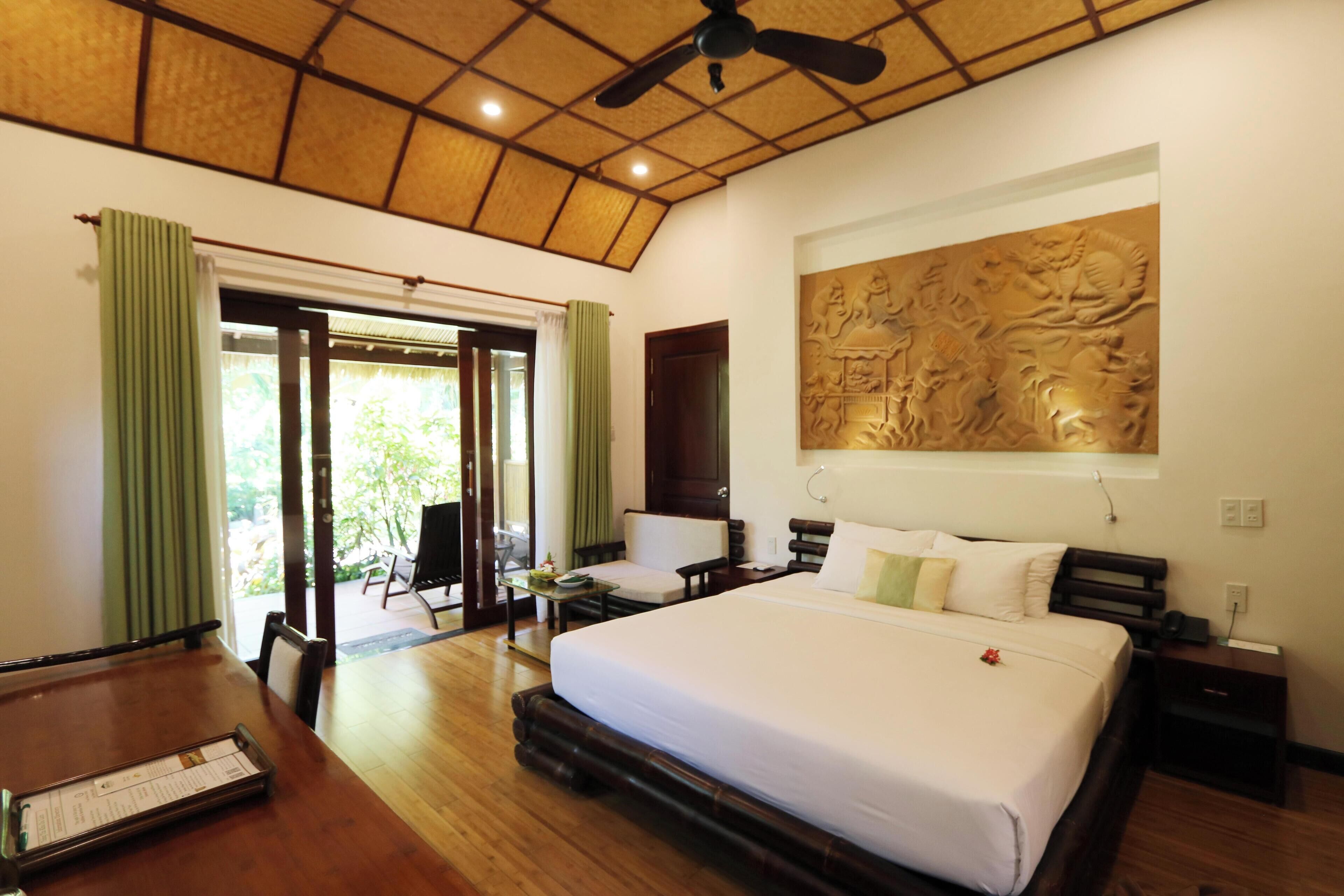 A hotel room with a large bed, a dining table, a ceiling fan, and a view of the outdoors.