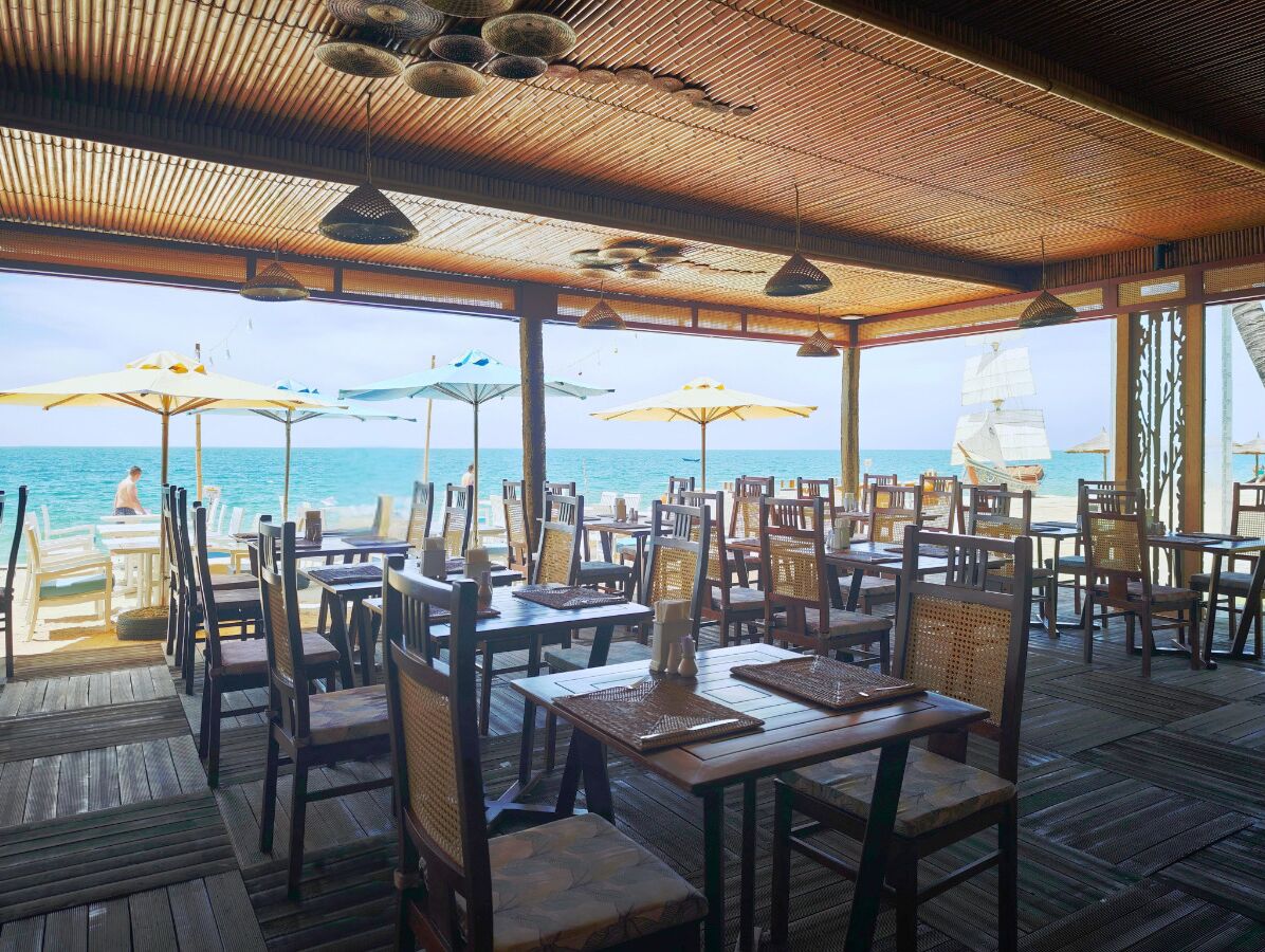 Un espacio de comedor al aire libre con mesas y sillas de madera, sombrillas y vista al mar.